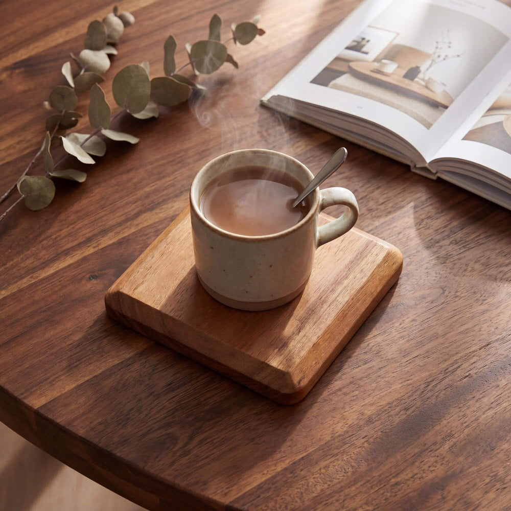 Styled tabletop setting featuring an acacia wood coaster beneath a ceramic mug on a wooden table.