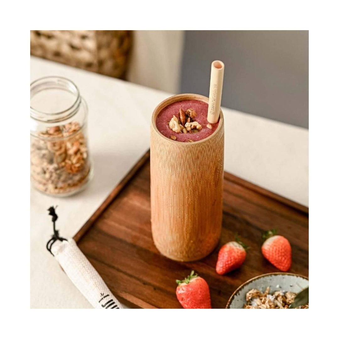 Bamboo cup filled with berry smoothie on wooden tray with strawberries and granola.