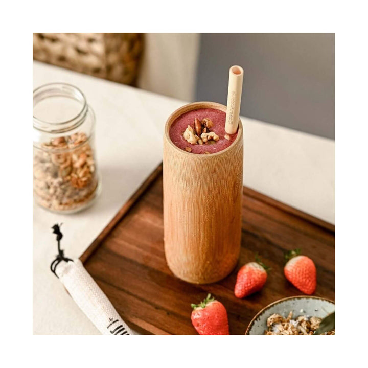 Bamboo cup filled with berry smoothie on wooden tray with strawberries and granola.