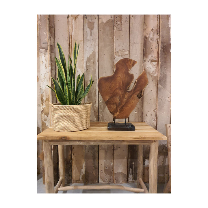 Teak figure stand displayed on a small teak table next to a woven planter and plant against rustic wooden wall.