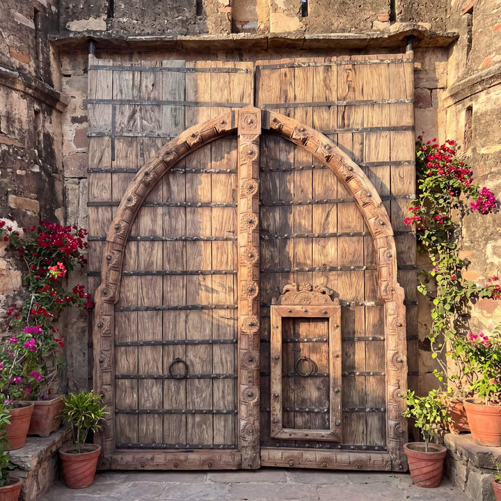 A large carved wooden double castle door with iron studs and a small inset door, set in a stone wall with potted flowers on both sides.