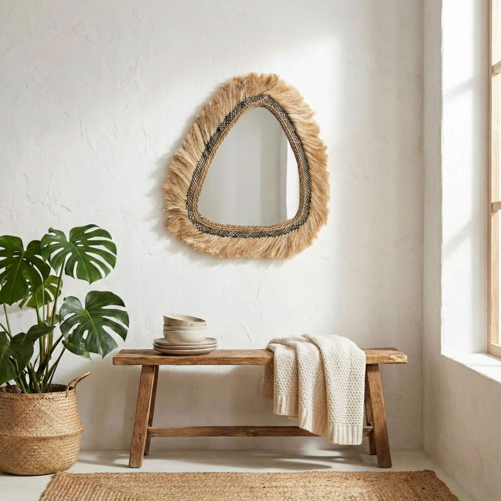 Woven Gemilang mirror above a wooden bench with a plant and rug in a bright room.