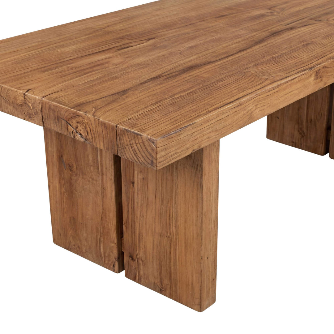 Angled view of the dining table highlighting its thick teak tabletop and bold legs.