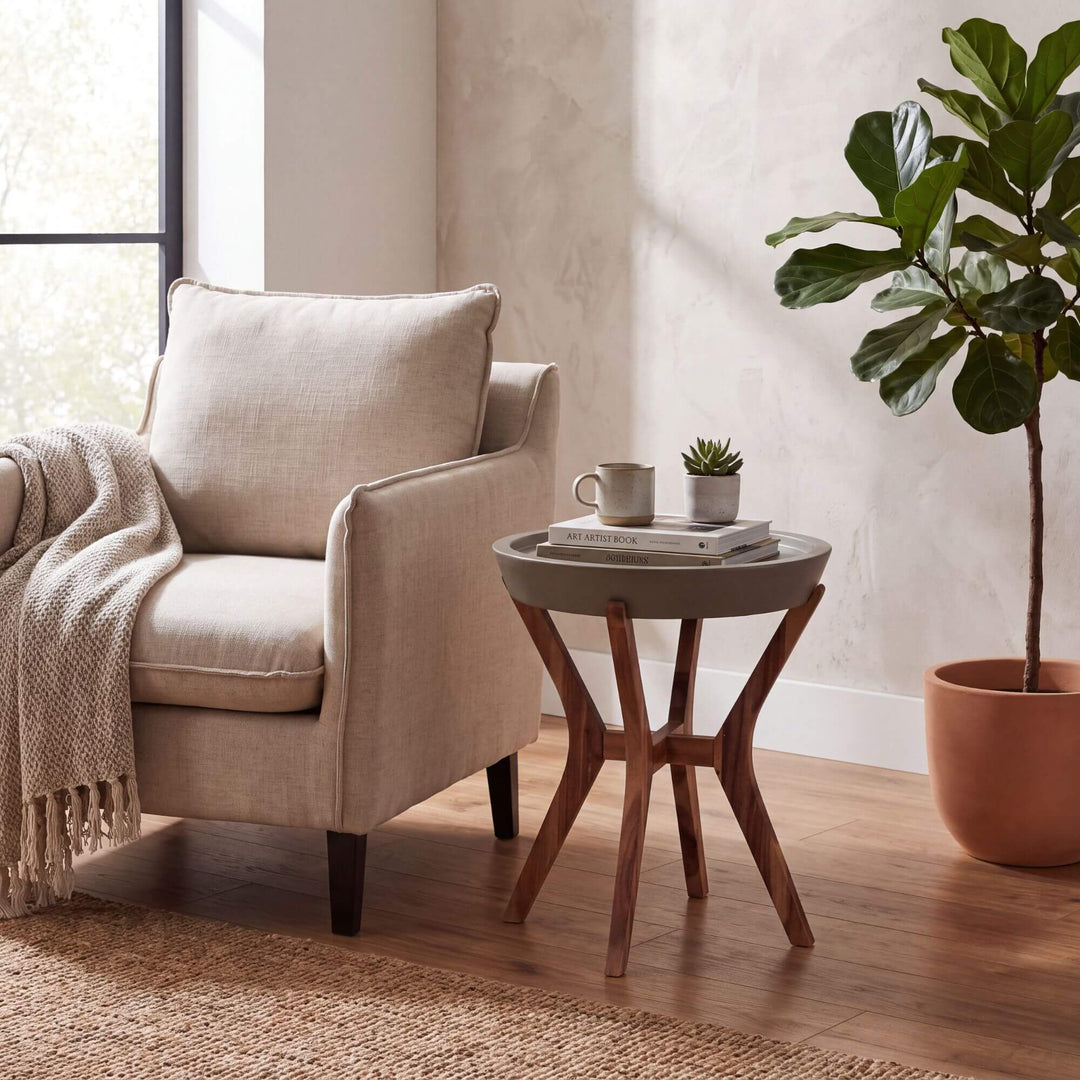 A beige upholstered armchair beside a round gray Kaia side table with a wooden star-shaped base, styled in a bright living room with a potted fiddle-leaf fig, books, a mug, and a throw blanket.