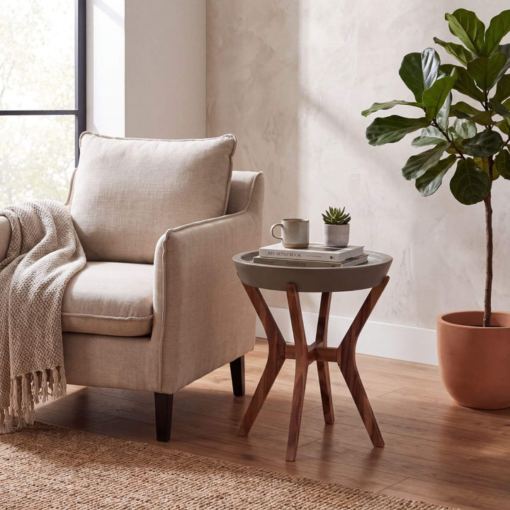 A beige upholstered armchair beside a round gray Kaia side table with a wooden star-shaped base, styled in a bright living room with a potted fiddle-leaf fig, books, a mug, and a throw blanket.