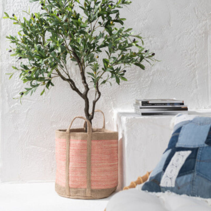 Potted plant in a KUTA JUTE BASKET next to a textured wall and a white surface with books and a pillow.