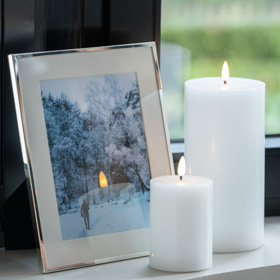Two Led white candles and a framed photo of a snowy landscape on a surface.