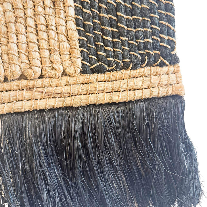 Close-up of Merak Wall Deco handwoven from abaca, rattan, and cotton with natural and black patchwork design and fringe