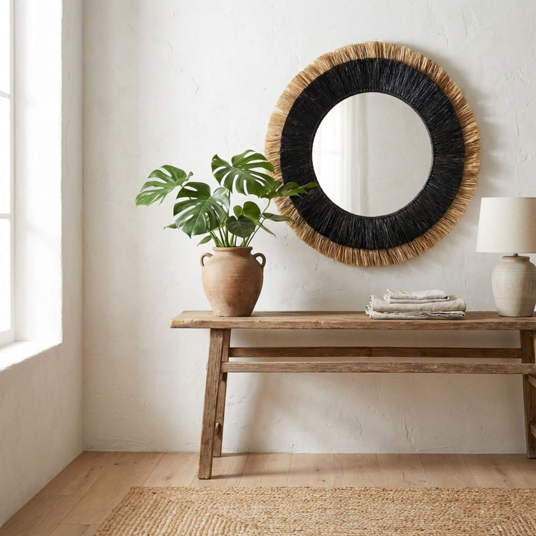 Round Nusa mirror with black frame and woven details above a wooden console table with a plant and lamp.