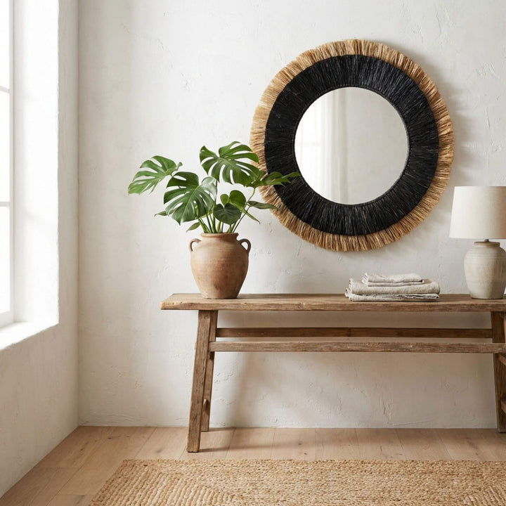 Round Nusa mirror with black frame and woven details above a wooden console table with a plant and lamp.