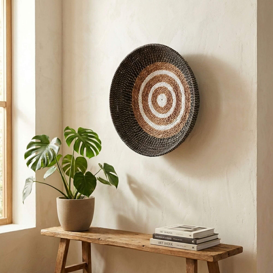 Woven Samara wall art piece on a beige wall with a plant and books on a wooden bench.