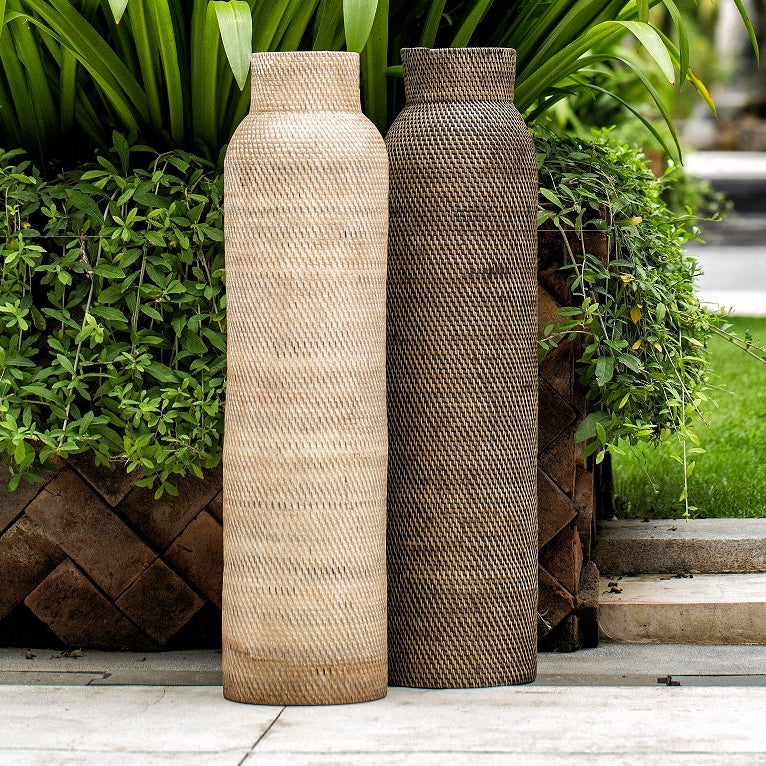 Two cylindrical wicker vases placed on a flat surface with green plants in the background, one vase is in focus and appears beige in color, while the other is blurred in the background.